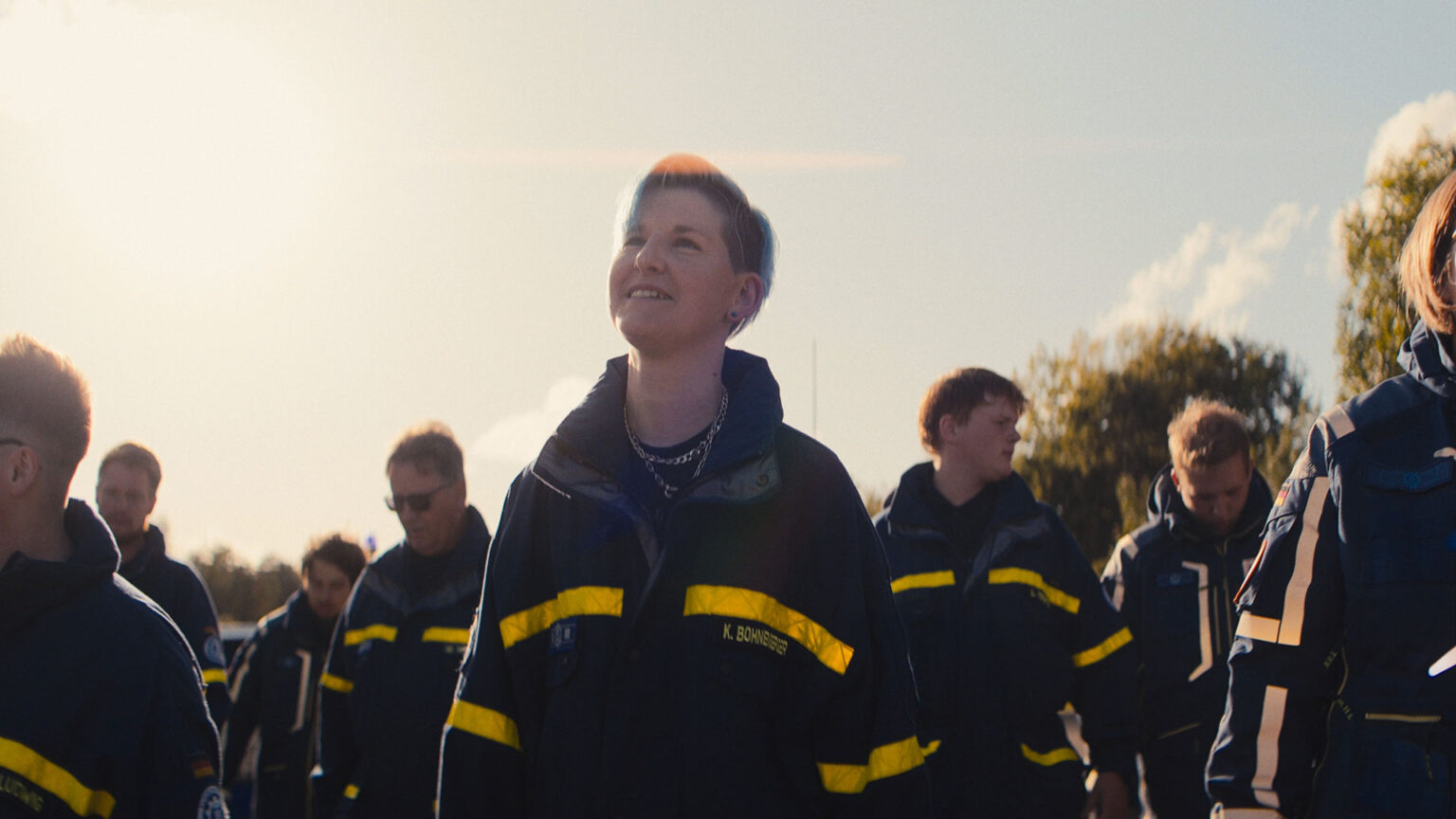 Eine Gruppe von Menschen in dunklen Uniformen mit gelben Streifen geht während eines Imagefilms zum Tag der deutschen Einheit im Freien im Sonnenlicht spazieren. Die Person in der Mitte lächelt, während sie nach oben schaut, mit Bäumen und einem hellen Himmel im Hintergrund.