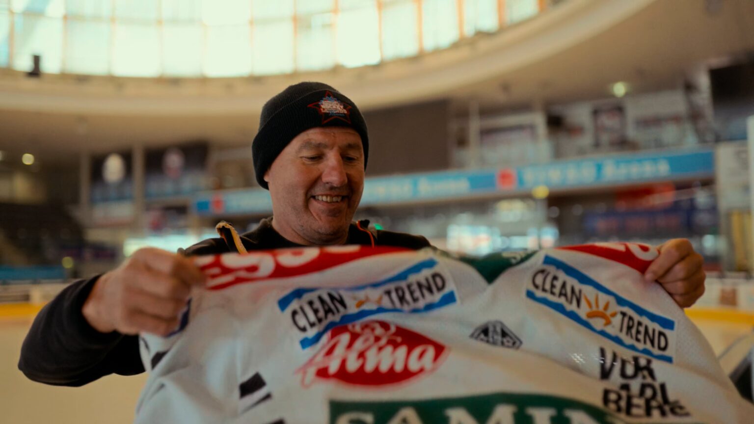 Ein lächelnder Mann mit einer Mütze hält ein weißes Sporttrikot hoch, das mit verschiedenen Sponsorenlogos bedruckt ist, während er in einer Eishalle mit großen Fenstern steht.